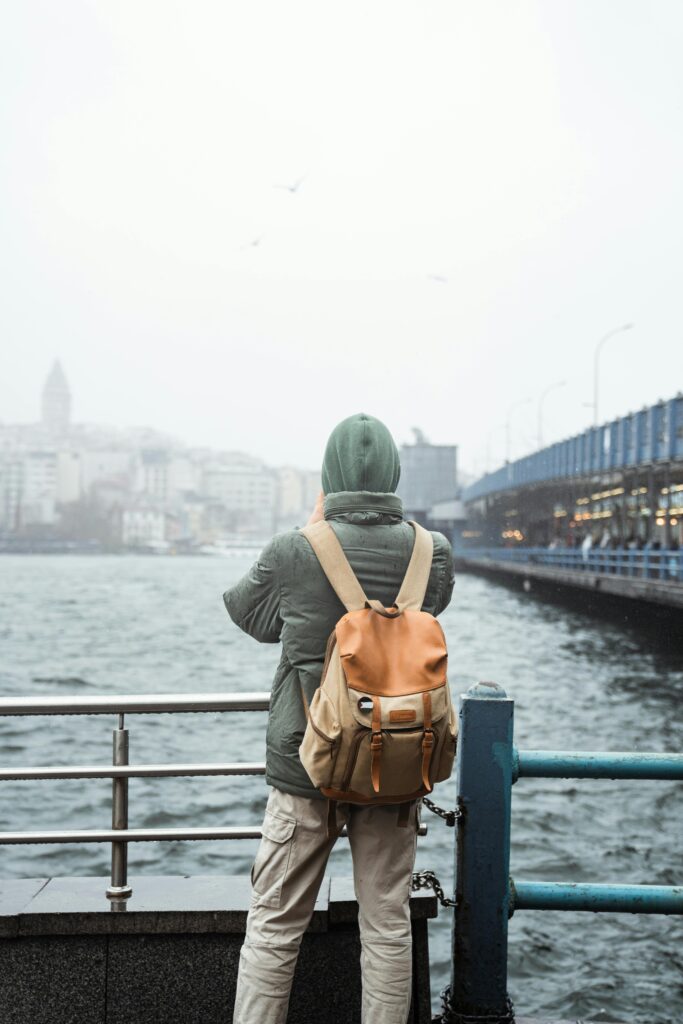 A traveler stands by a riverside railing, gazing at an urban skyline on a cloudy day.